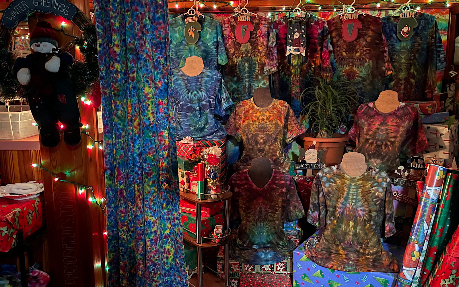 Just to the right of the tie dye bar, are five t-shirt mannequins each wearing a new, symmetrical psychedelic mindscape ice dye design. The entire frame is full of Christmas decorations, gift wrapped boxes, and ambient red and green lights strung about