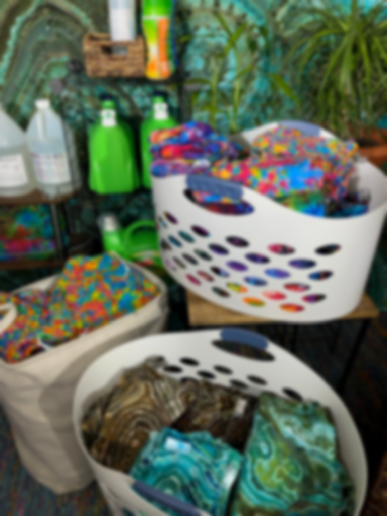 Three laundry baskets full of folded tie dye, with jugs of detergent and bottles of Dharma Trading Co. Synthrapol on shelves in the background
