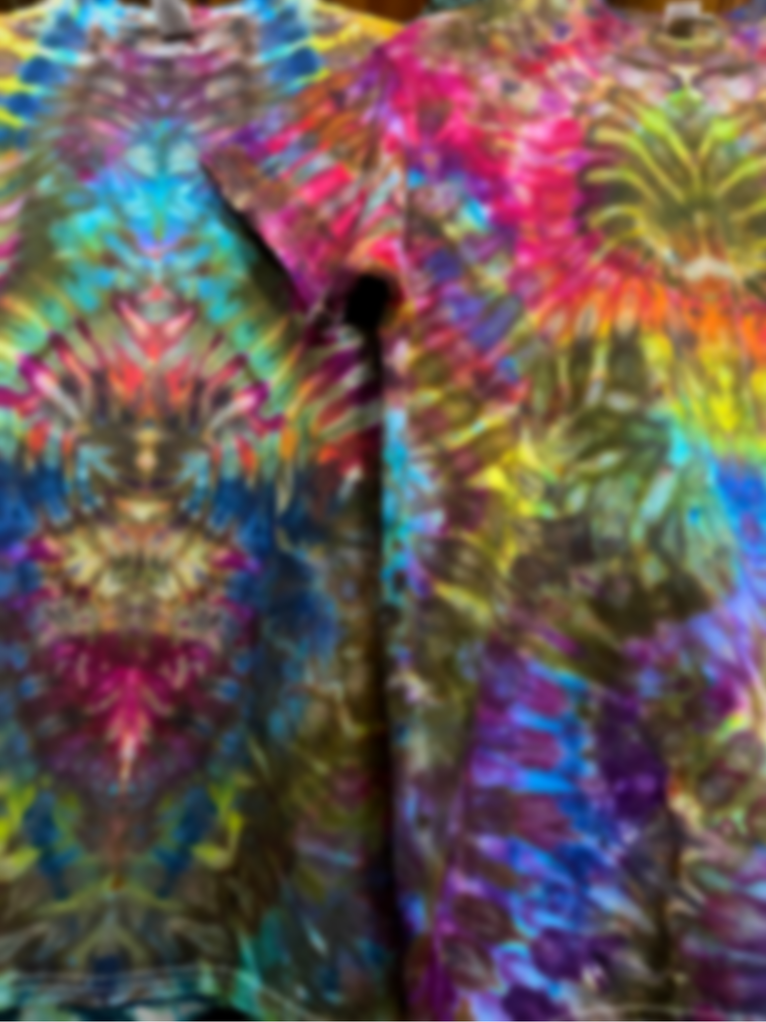 A slightly out of focus, close up shot of two rainbow psychedelic mindscape symmetrical ice dyed t-shirts hanging next to one another