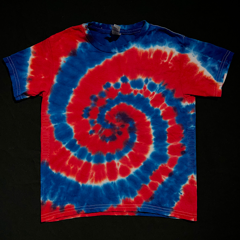A toddler sized short sleeve t-shirt featuring a Fourth of July red, white & blue spiral tie dye design; laid flat on a solid black background 