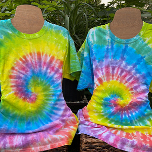 Two retro rainbow pastel spiral tie dye t-shirts displayed on mannequins, outdoors in front of a luscious green tropical fig tree