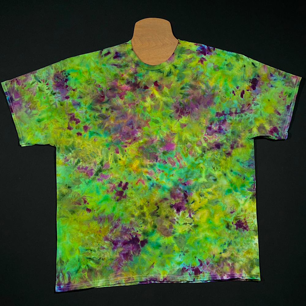 An example of a finished, made-to-order Purple Urkle ice dyed t-shirt, a splatter tie dye pattern featuring an array of green and purple shades, inspired by the appearance of a marijuana bud, laid flat on a solid black background
